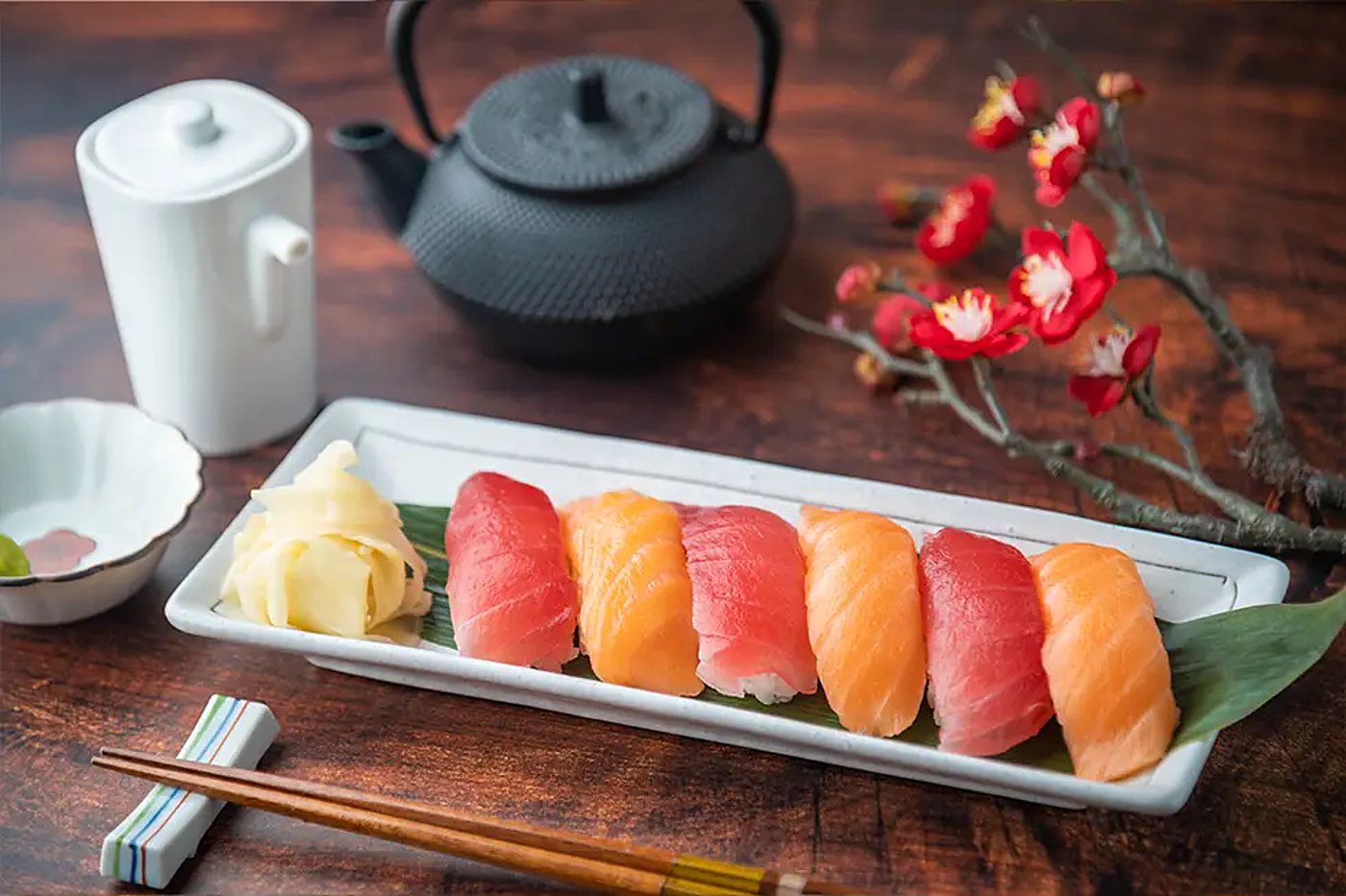 Une assiette rectangulaire de six nigiris au thon et au saumon avec du gingembre mariné, posée sur une table en bois près d'une théière en fonte et d'une branche de fleurs rouges.