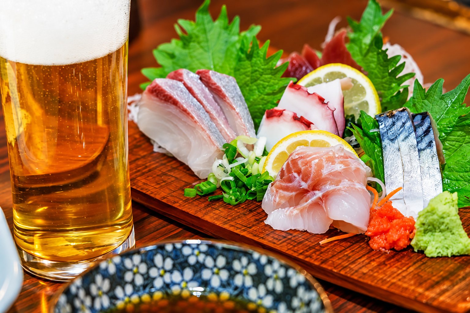 Une planche en bois garnie de tranches de poisson cru, de feuilles de shiso, de citron et de wasabi, à côté d'un grand verre de bière blonde.