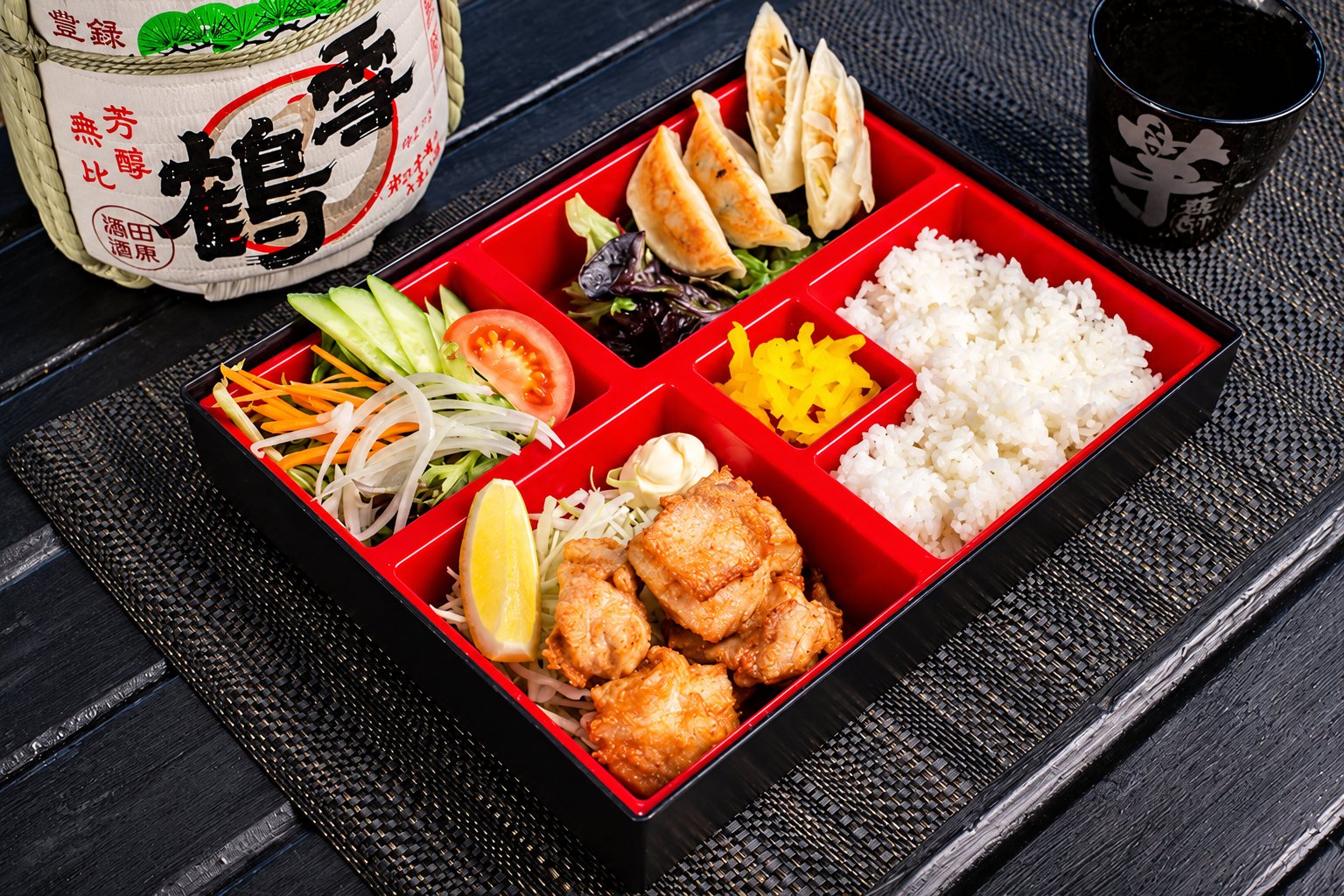 Un bento laqué rouge et noir contenant du poulet frit, des gyozas, du riz blanc, des pickles jaunes et une salade, posé sur un set de table devant un tonneau de saké.