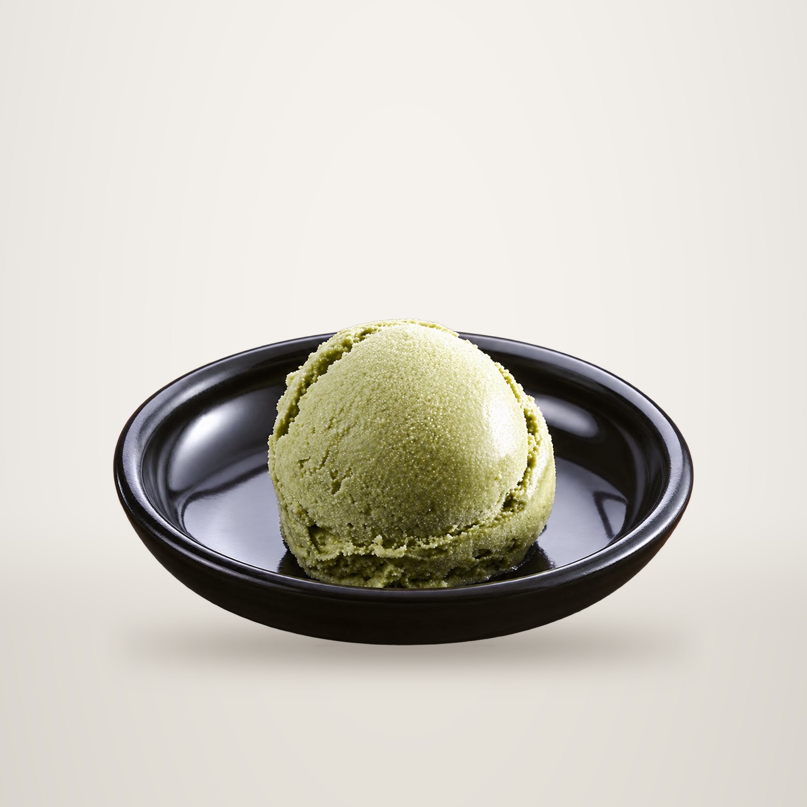 Une boule de glace de couleur vert pâle, probablement au thé vert, posée dans une petite coupelle sombre sur fond blanc.