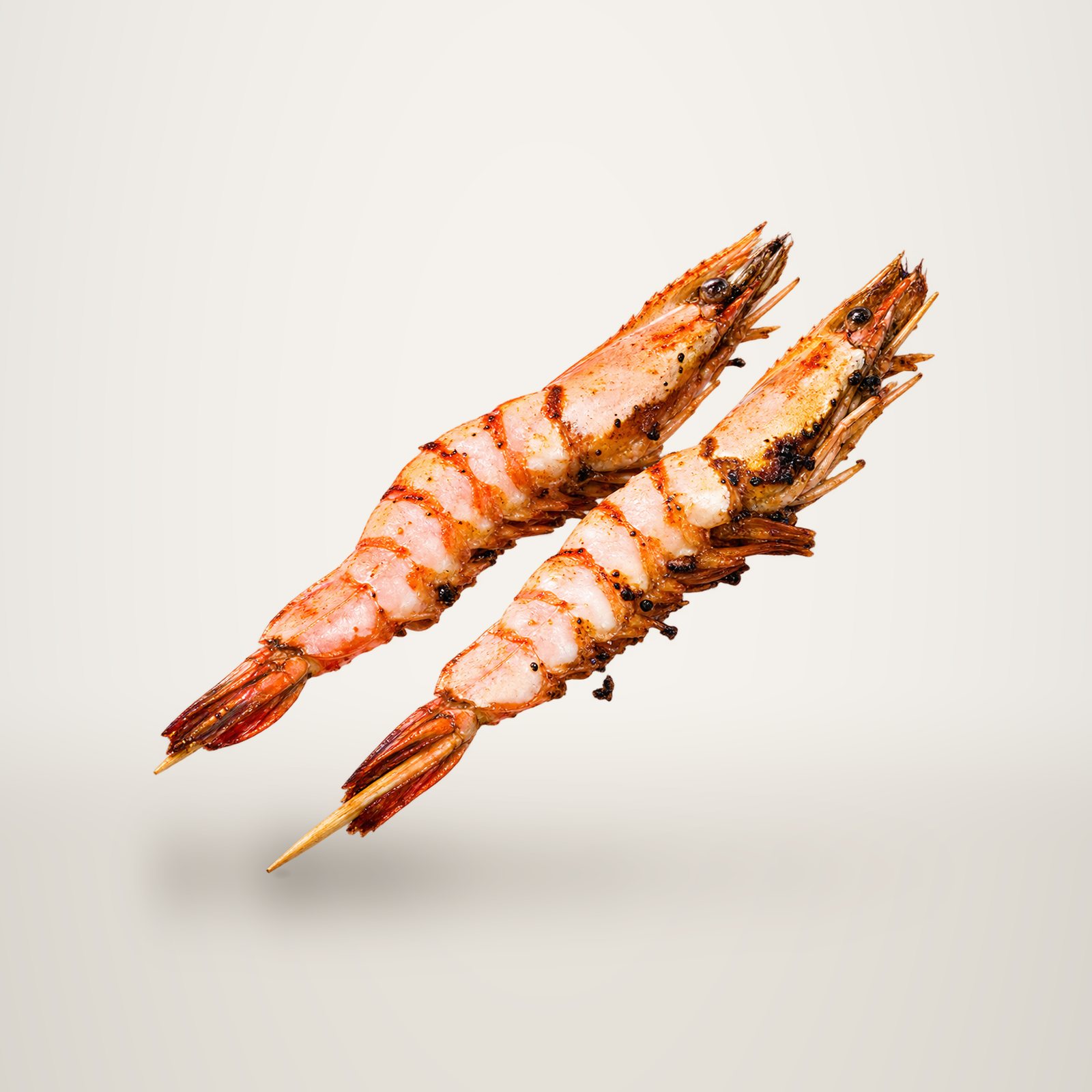Deux grosses crevettes décortiquées et grillées, enfilées sur des piques en bois, photographiées sur fond blanc.