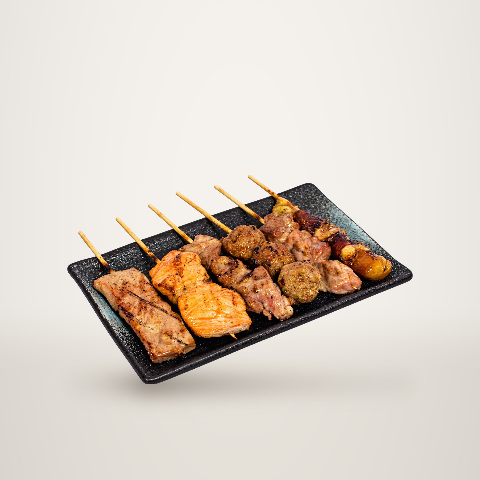Plateau noir rectangulaire garni de brochettes grillées de saumon, de viandes et de légumes sur des piques en bambou, posé sur fond blanc uni.