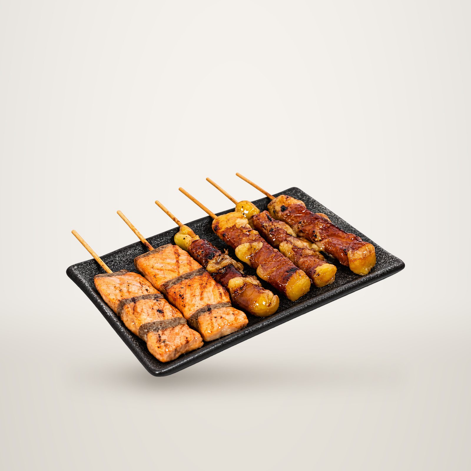 Un plateau rectangulaire noir présente six brochettes grillées et laquées, dont deux de saumon et quatre de viande enroulée autour de morceaux dorés, sur fond blanc.