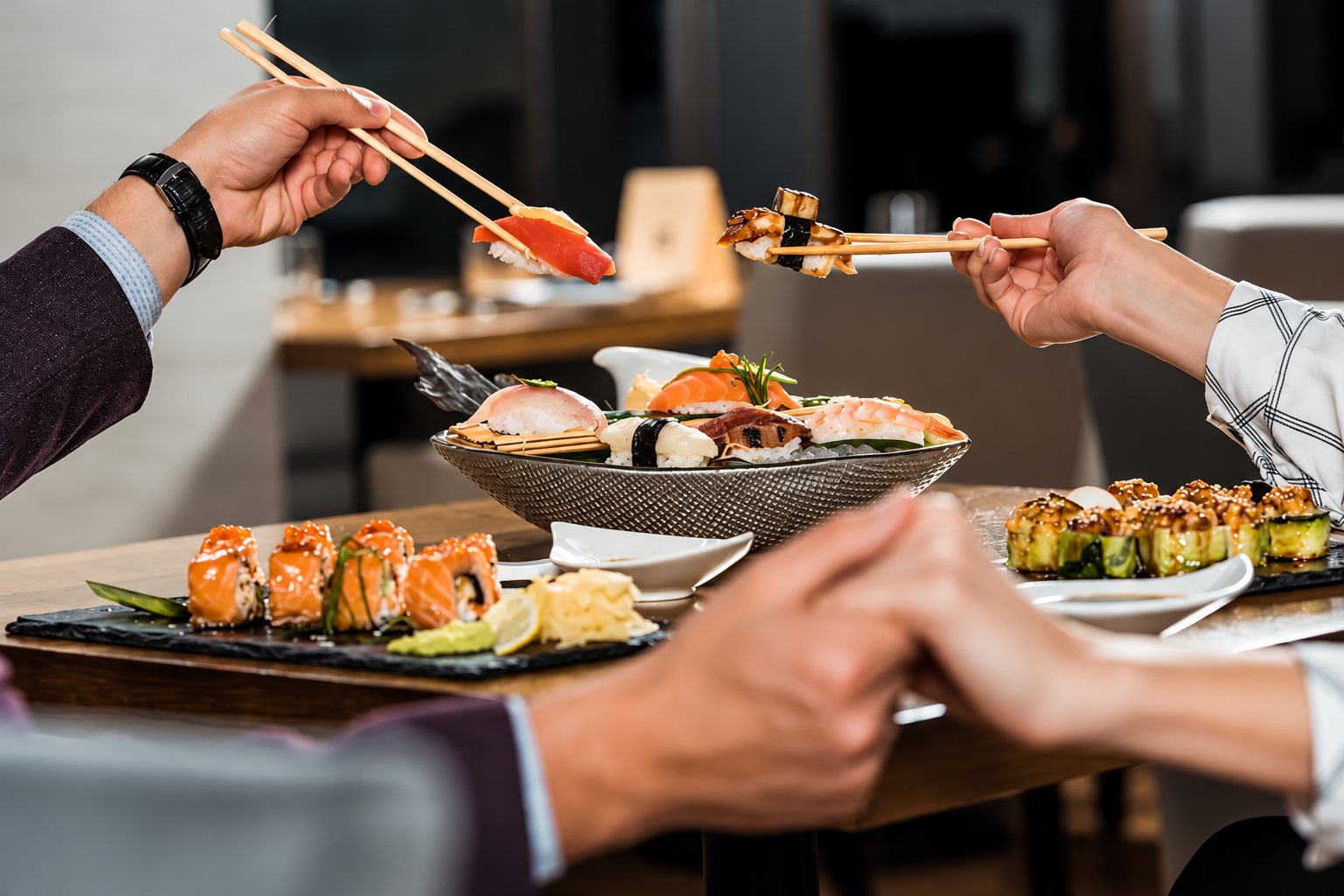 Deux personnes se tenant la main tendent leurs baguettes vers des assiettes de sushis et de makis posées sur une table de restaurant.