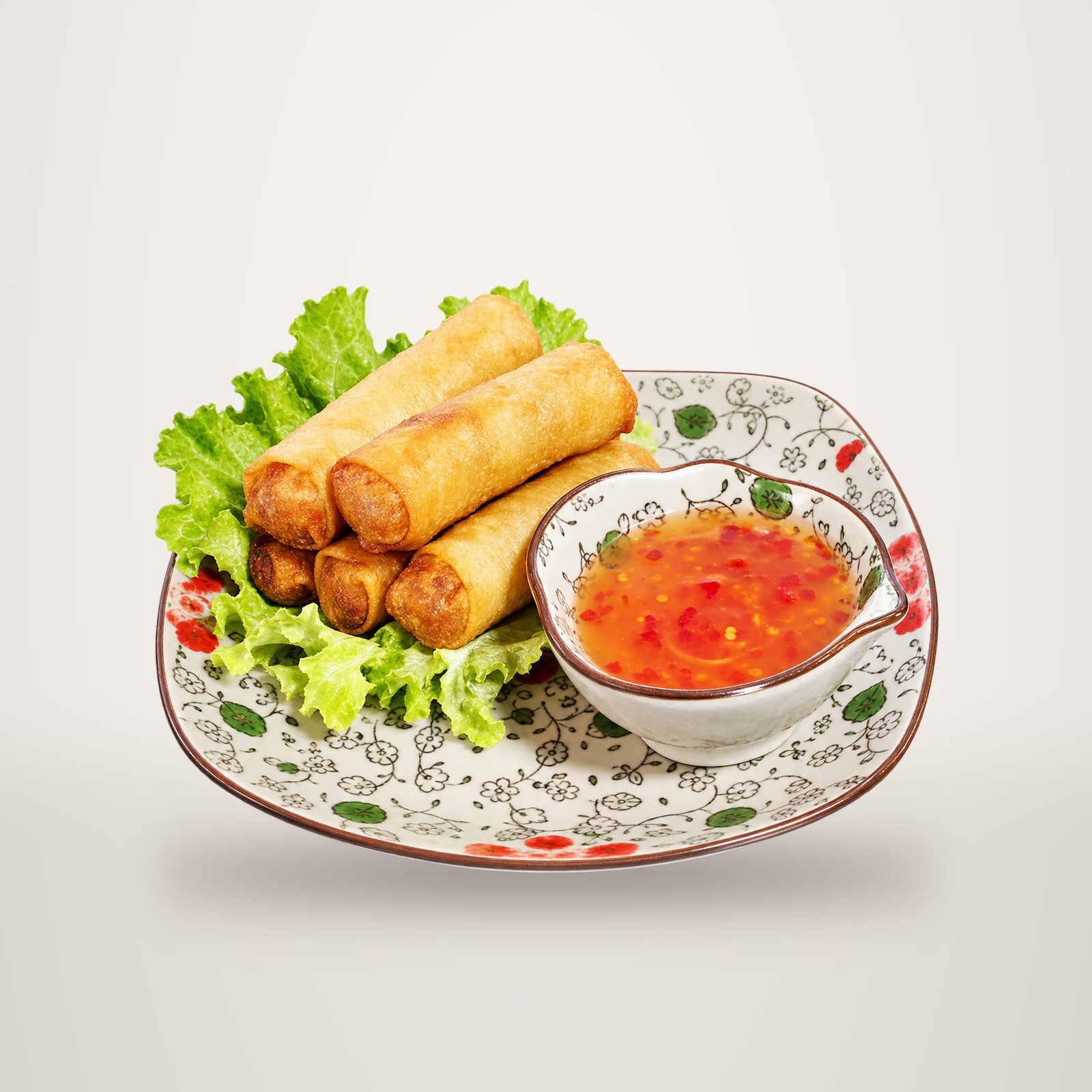 Des nems dorés sont servis sur des feuilles de salade verte dans une assiette décorée, accompagnés d'un petit bol de sauce rouge.