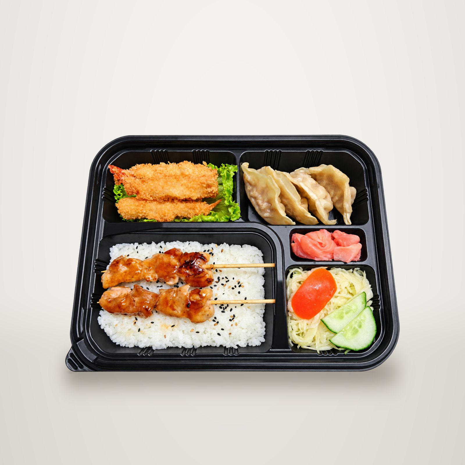 Vue de dessus d'un plateau bento noir contenant du riz au sésame avec deux brochettes de poulet, des crevettes panées, des gyozas, du gingembre mariné et une salade de chou avec concombre et tomate.