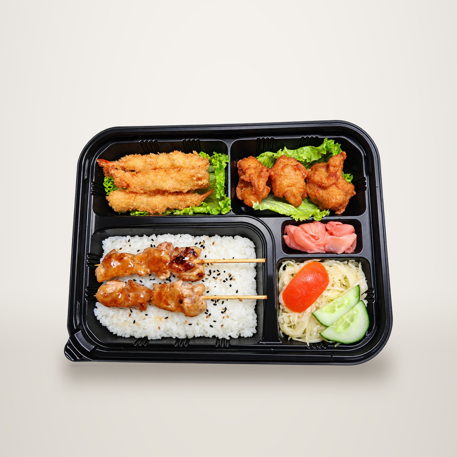 Vue de dessus d'un plateau repas noir à compartiments contenant du riz au sésame avec deux brochettes de poulet yakitori, des crevettes panées, des morceaux de poulet frit, du gingembre mariné et une salade.