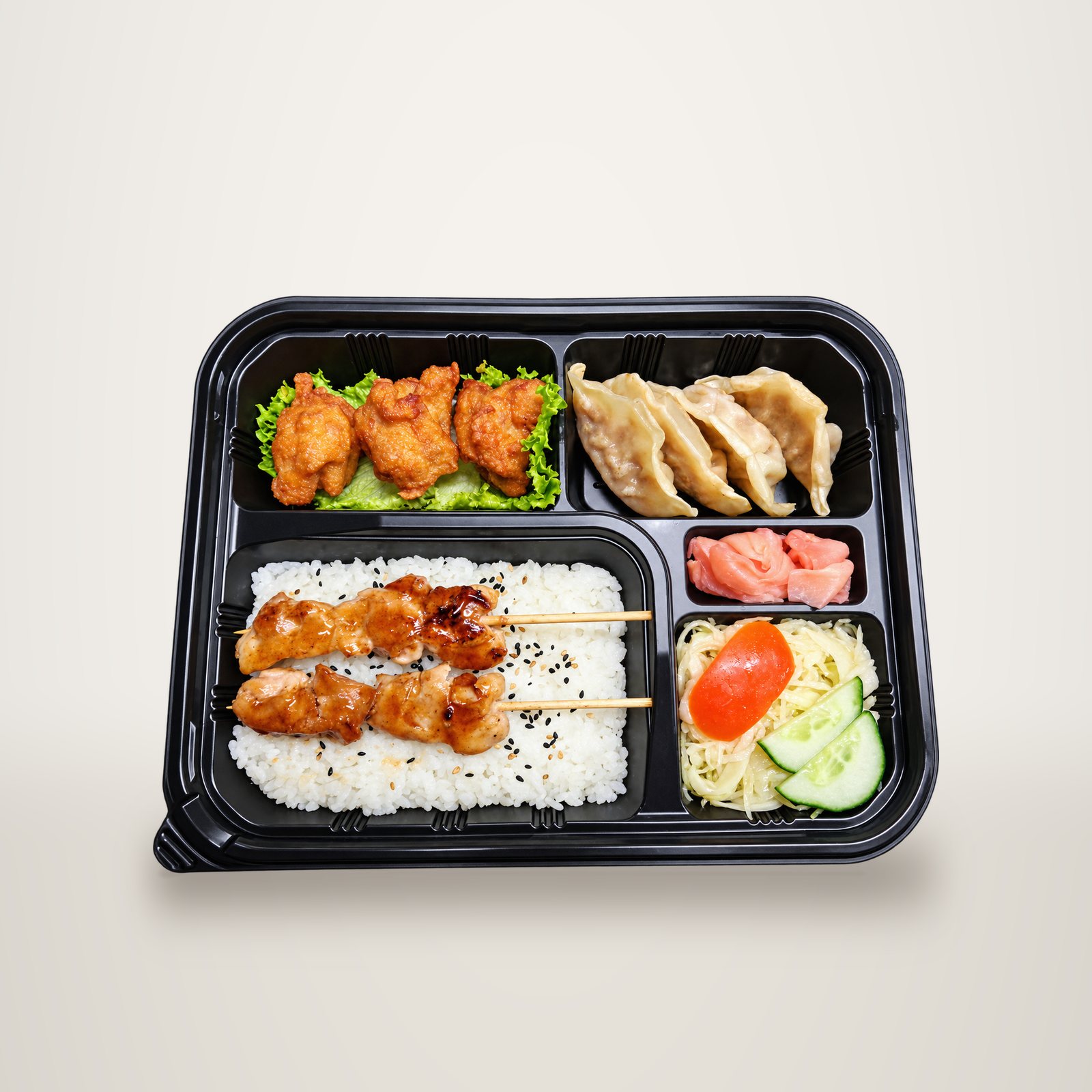 Vue de dessus d'un plateau-repas noir contenant du riz au sésame avec deux brochettes de poulet teriyaki, des beignets de poulet, des gyozas, du gingembre mariné et une salade de chou avec concombre.