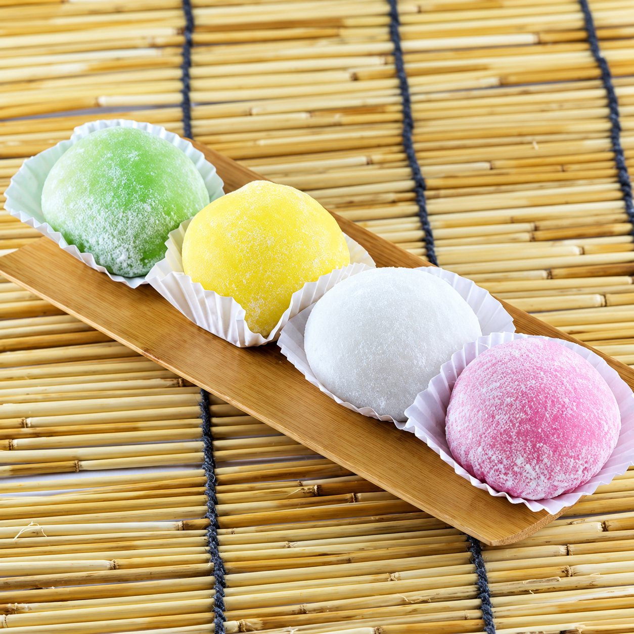 Quatre mochis vert, jaune, blanc et rose présentés dans des caissettes en papier sur un plateau en bois posé sur un tapis de bambou.
