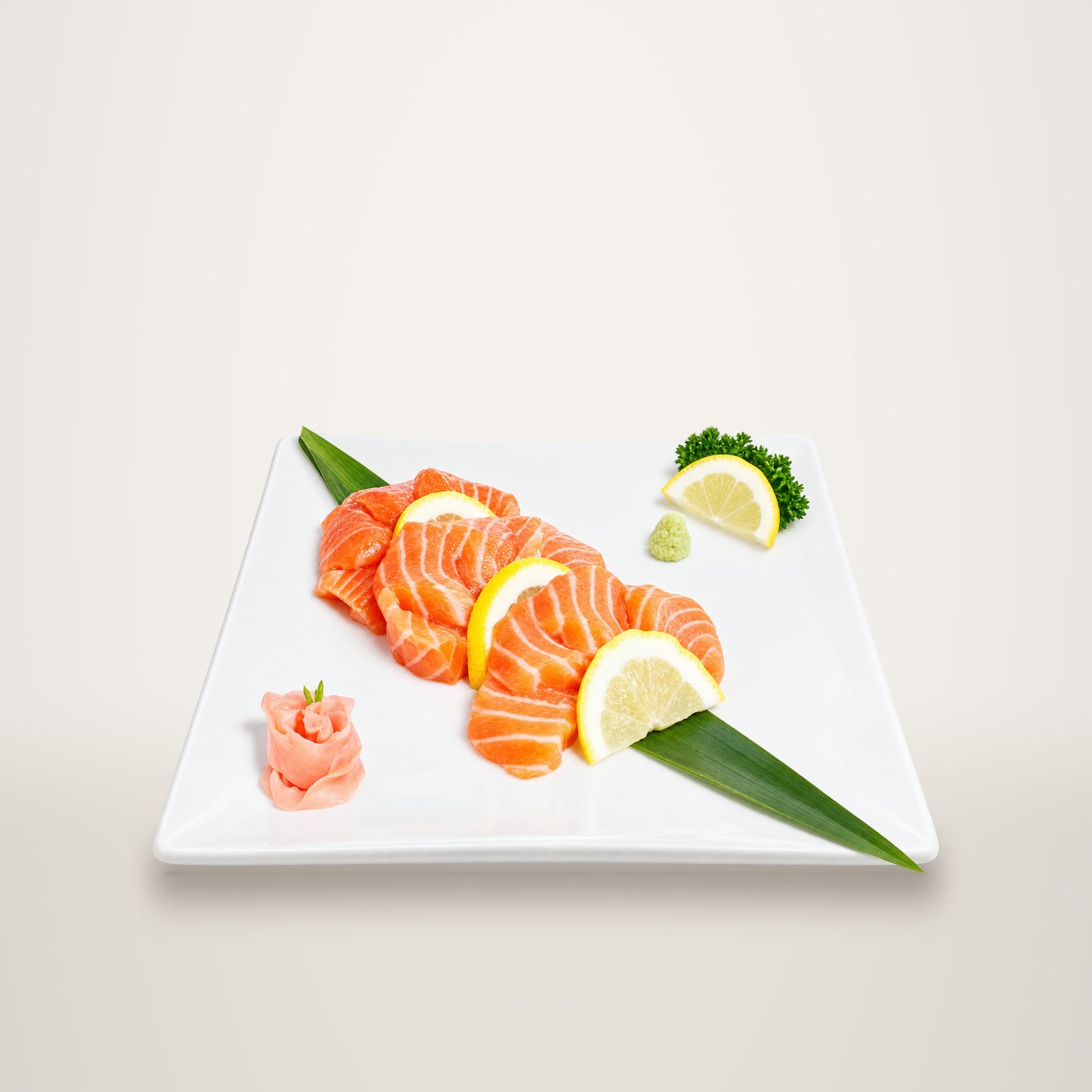 Tranches de sashimi de saumon posées sur une feuille de bambou, avec quartiers de citron, gingembre mariné et wasabi sur une assiette blanche.